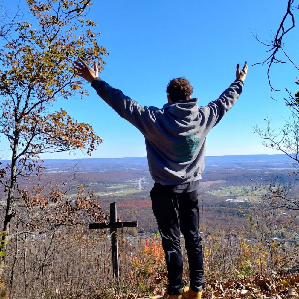 A man standing on a mountain overlook beside a wooden cross, arms raised toward the valley below